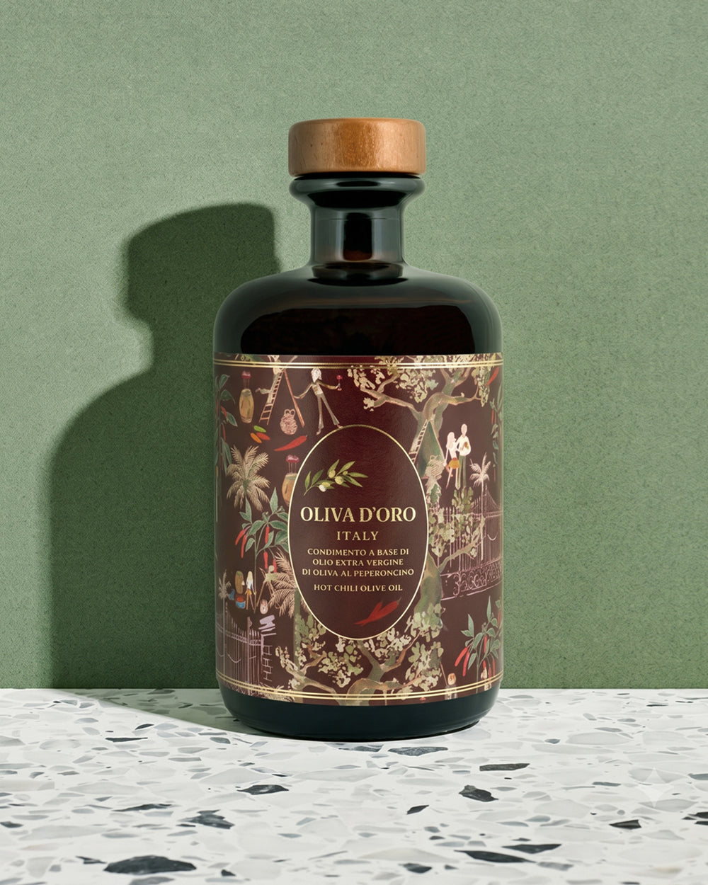 A dark glass bottle of Oliva D'Oro olive oil with a wooden cap, set against a textured sage green background and a white speckled surface. Available via the Pantry Collection from Wildschut Antiques.