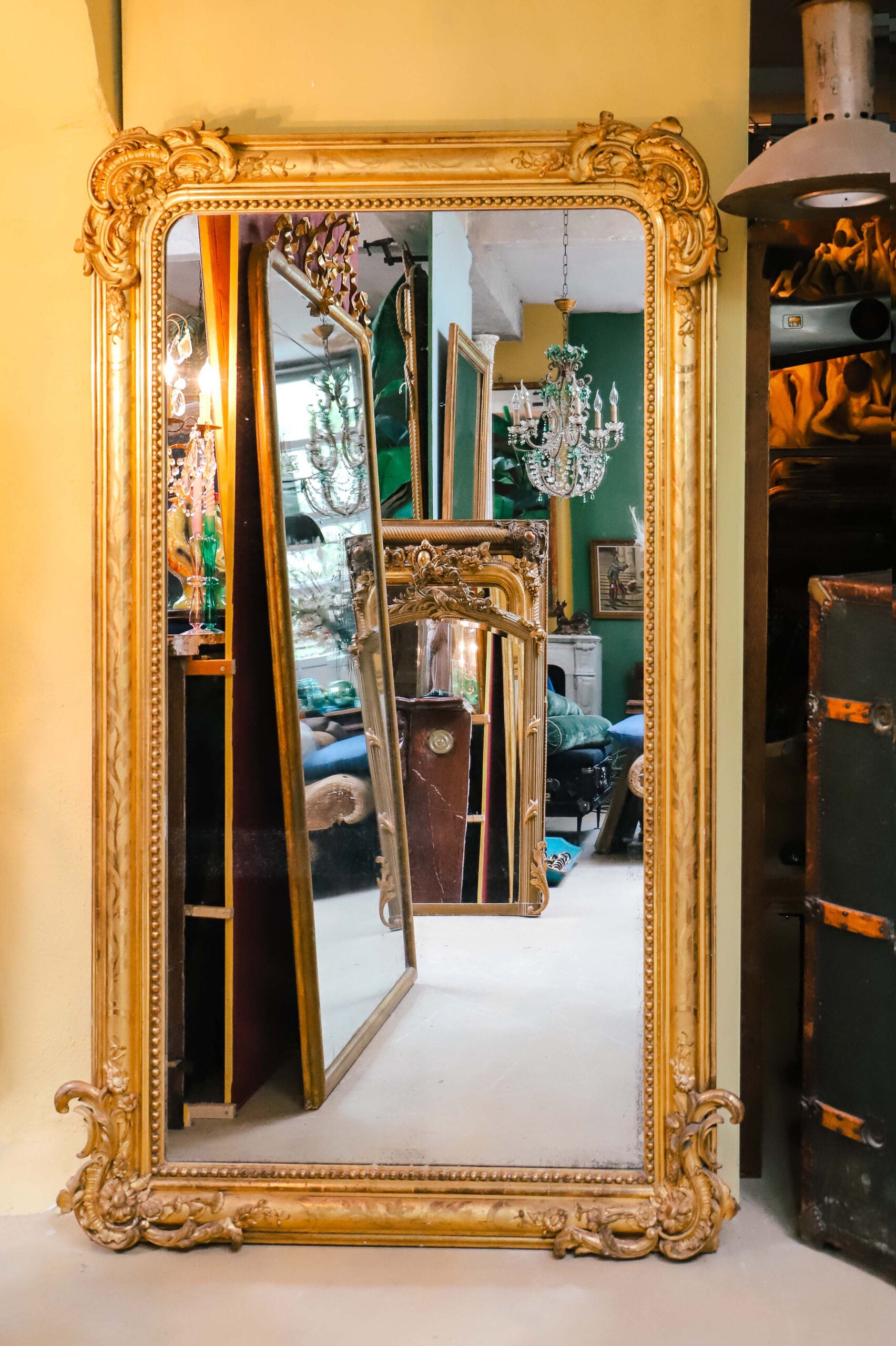 Large Antique French 19th Century gold gilt mirror with leaf and flower ornaments in the frame, displayed in the warehouse of Wildschut Antiques against a yellow backdrop and reflecting other mirrors and antique furniture pieces