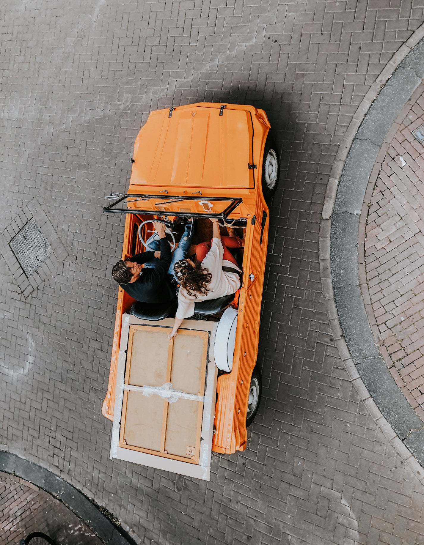 Overhead shot of Baukje and Michiel Wildschut cruising through the streets of Amsterdam in their orange Citroën Méhari, holding onto a painting in the open back—en route for delivery.