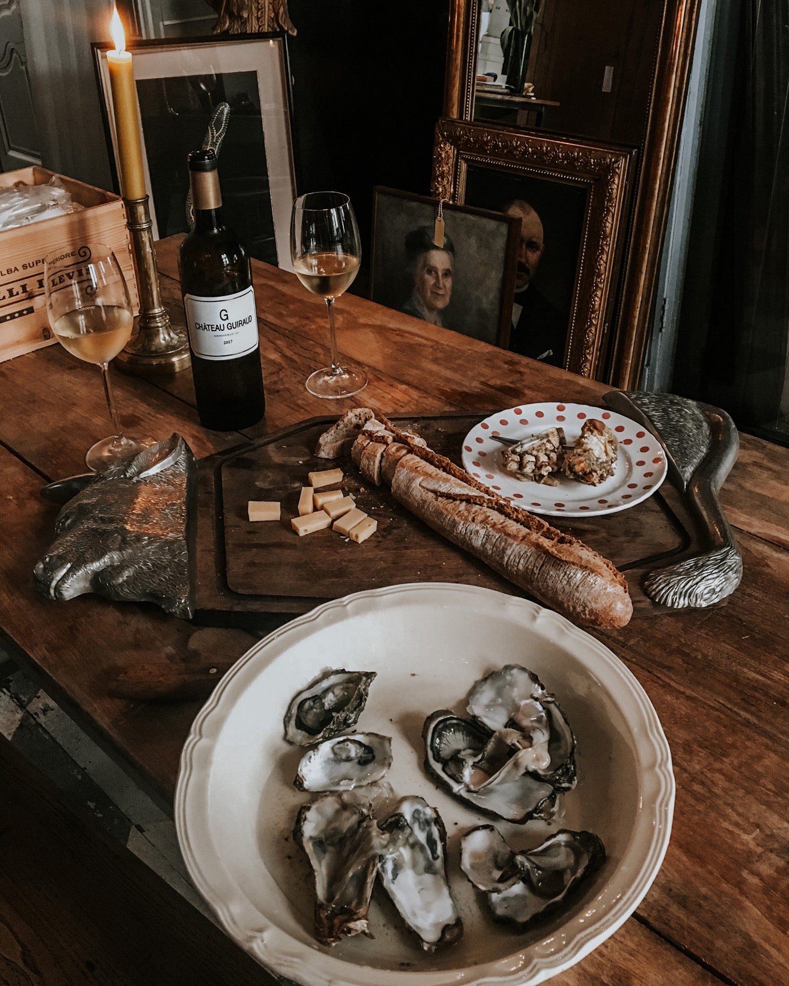 A classical table setting on a wooden table featuring a whimsical platter with a cow’s head and tail, French bread, cheese, and a platter of oysters. Two glasses of Château Guiraud wine stand nearby, with a yellow candle flickering in a brass holder. Antique portrait paintings provide a moody backdrop—all bathed in soft, dreamy light at Wildschut Antiques.