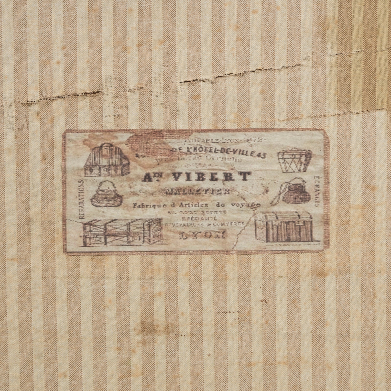 Close-up of the interior of a brown vintage canvas cube-shaped trunk, lined with striped linen fabric and featuring the maker’s label: A. Vibert, Lyon.