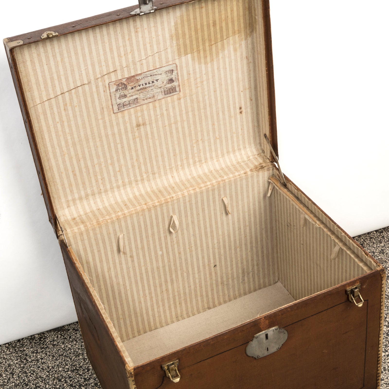 Interior view of a brown vintage canvas cube-shaped trunk, lined with striped linen fabric and featuring metal accents, and the maker’s label: A. Vibert, Lyon.