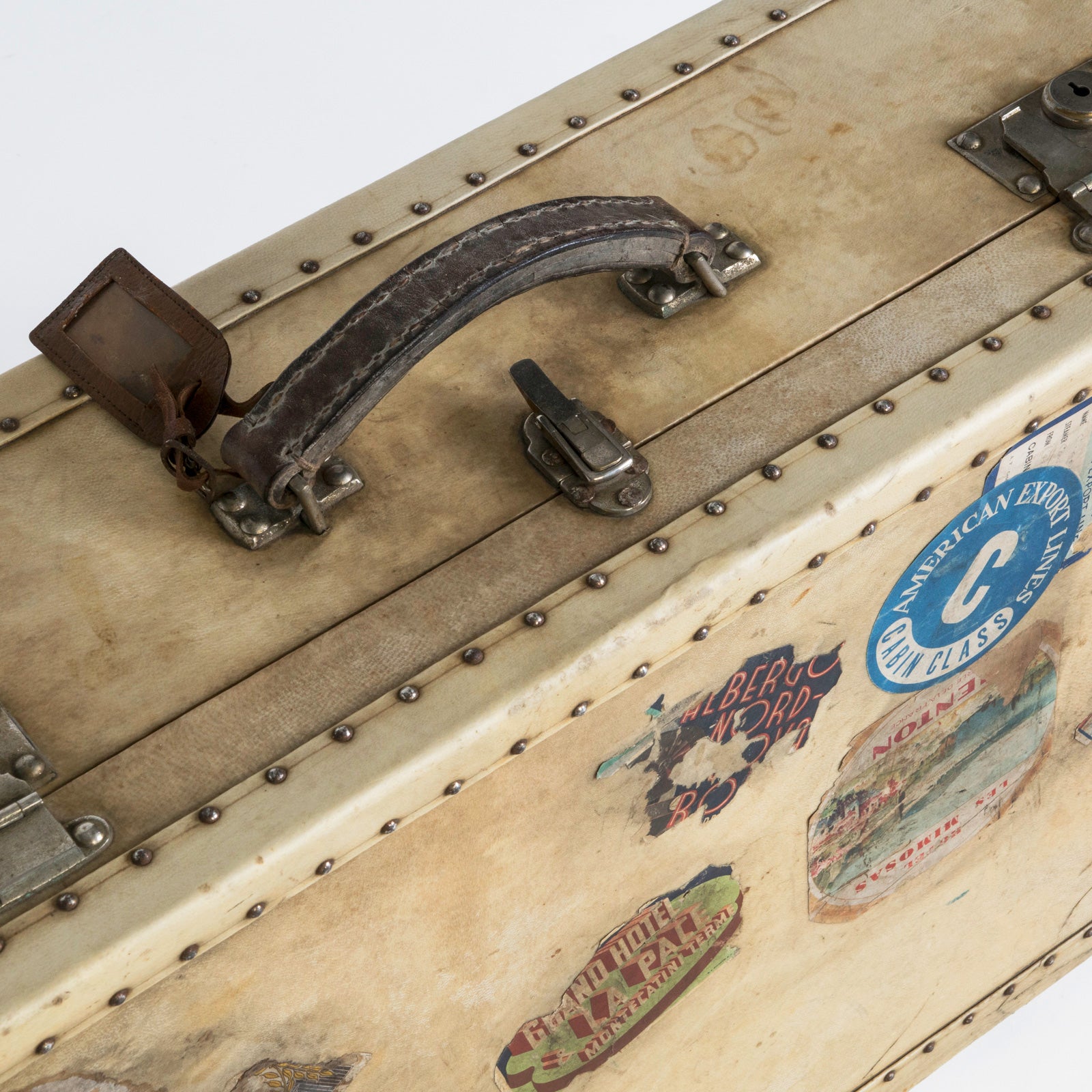 Close-up of the leather handle of a vintage French cream vellum white leather suitcase or valise with brass studs and lock plates, adorned with various travel labels and stickers