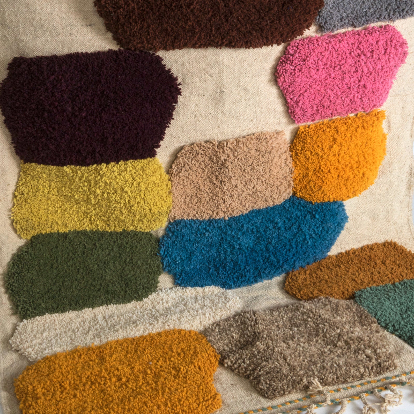 Close-up of a vintage 1970s Moroccan rug featuring an abstract composition of vibrant, irregularly shaped tufts in bold colors—such as cobalt blue, mustard yellow, fuchsia, burnt orange, and olive green—set against a natural cream background. The rug has a playful, textured appearance and is finished with fringed tassels on both ends.