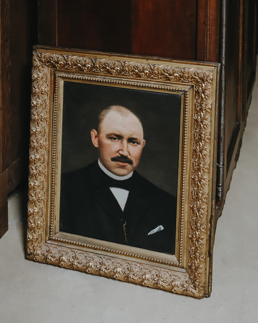 Antique oil portrait of a gentleman with a moustache, bald head, and dark suit, expertly painted by W.N. Hendrikse, displayed leaning against an antique wooden armoire in the warehouse of Wildschut Antiques