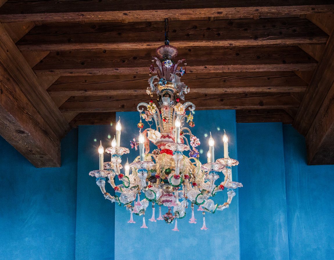 Magnificent and richly ornamented antique Venetian Murano glass chandelier with 12 lights, suspended from a wooden ceiling in an Italian palazzo with blue-toned walls.