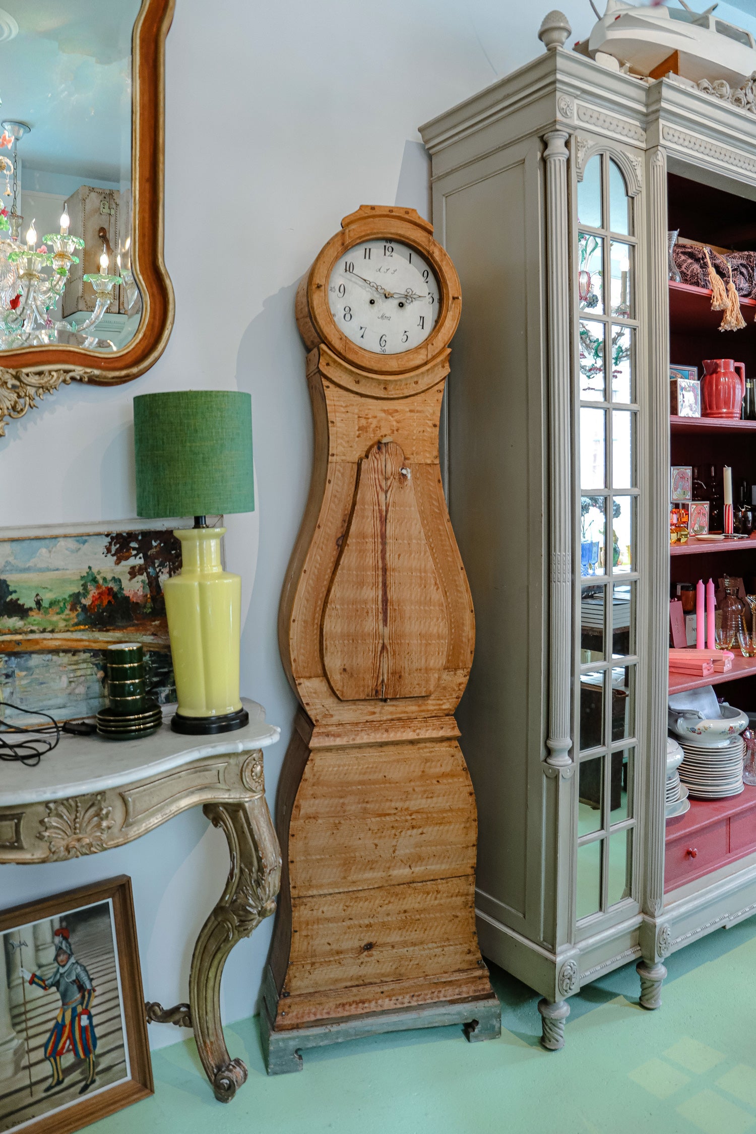 Antique Swedish Mora clock with a natural wood finish, wide belly shape, and numeral face, displayed beside a Louis XVI-style mirrored armoire and a console table topped with a yellow porcelain lamp with a green shade and a Rococo antique mirror—at Wildschut Antiques.
