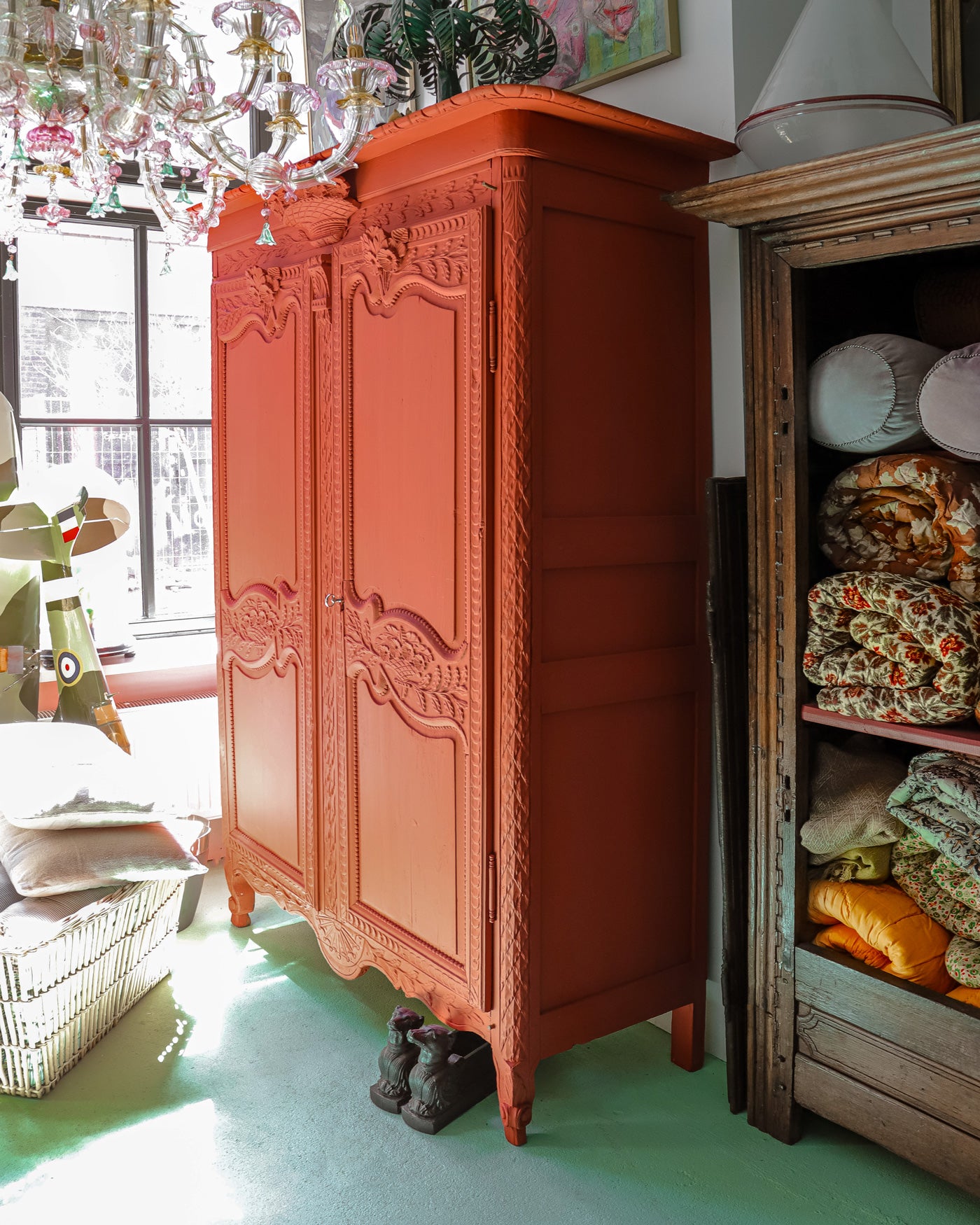 Side view of an antique French marriage armoire painted vibrant brick red with intricate carvings, positioned beside a dark oak armoire filled with vintage plaids and blankets. A pink Venetian glass chandelier hangs above—all at Wildschut Antiques.