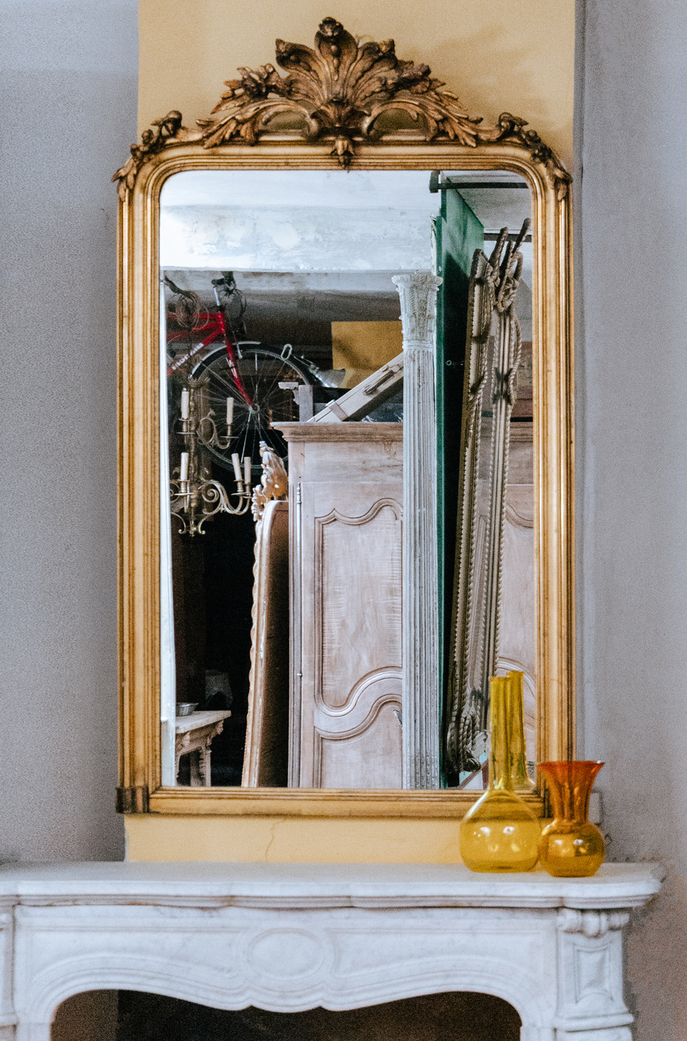 An antique gilded mirror rests atop a white marble fireplace adorned with yellow vases. The mirror reflects an antique armoire and architectural pillars in the warehouse of Wildschut Antiques.