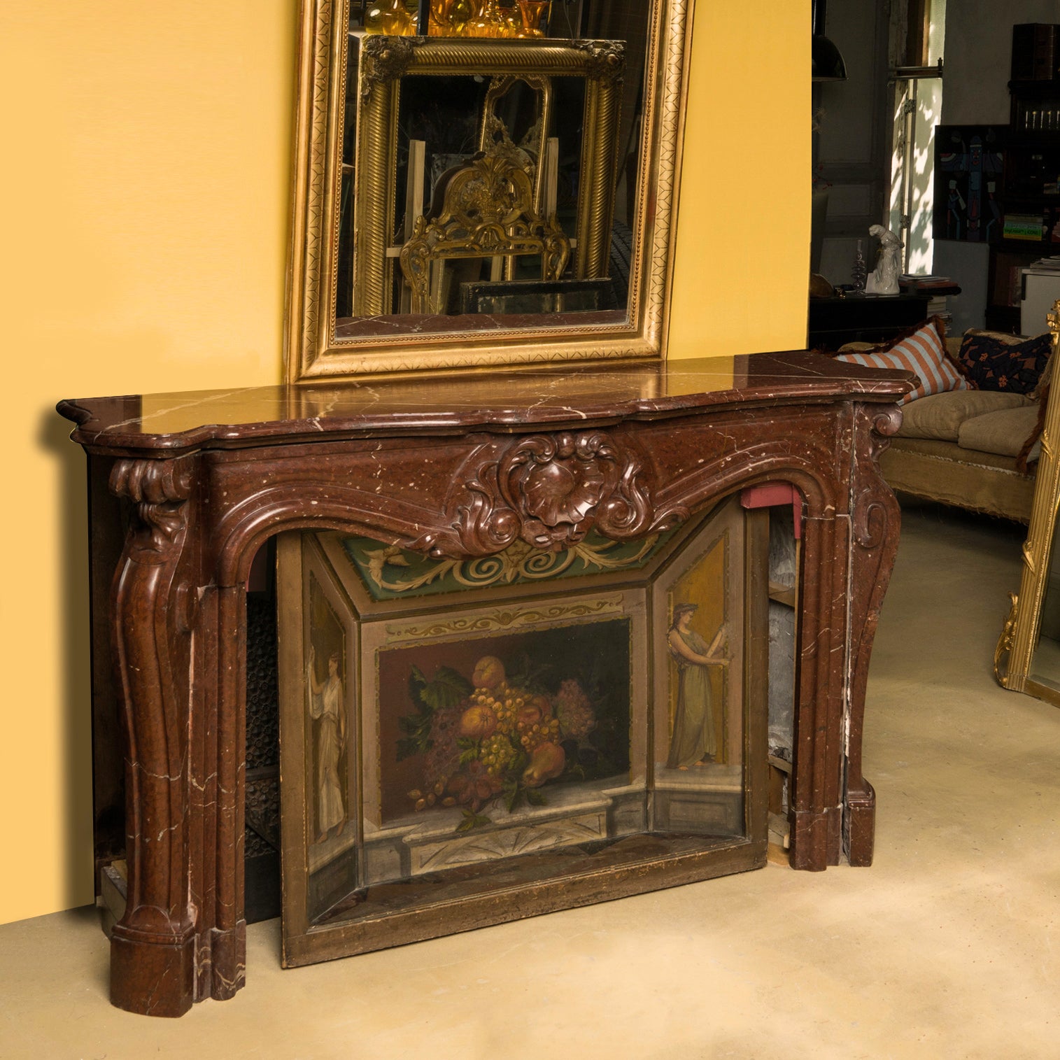 A grand antique French Rococo Louis XV fireplace mantel, crafted from richly veined red-brown marble. Displayed with an elegant painted firescreen adorned with fruits and two graceful female figures, all set in the atmospheric warehouse of Wildschut Antiques.