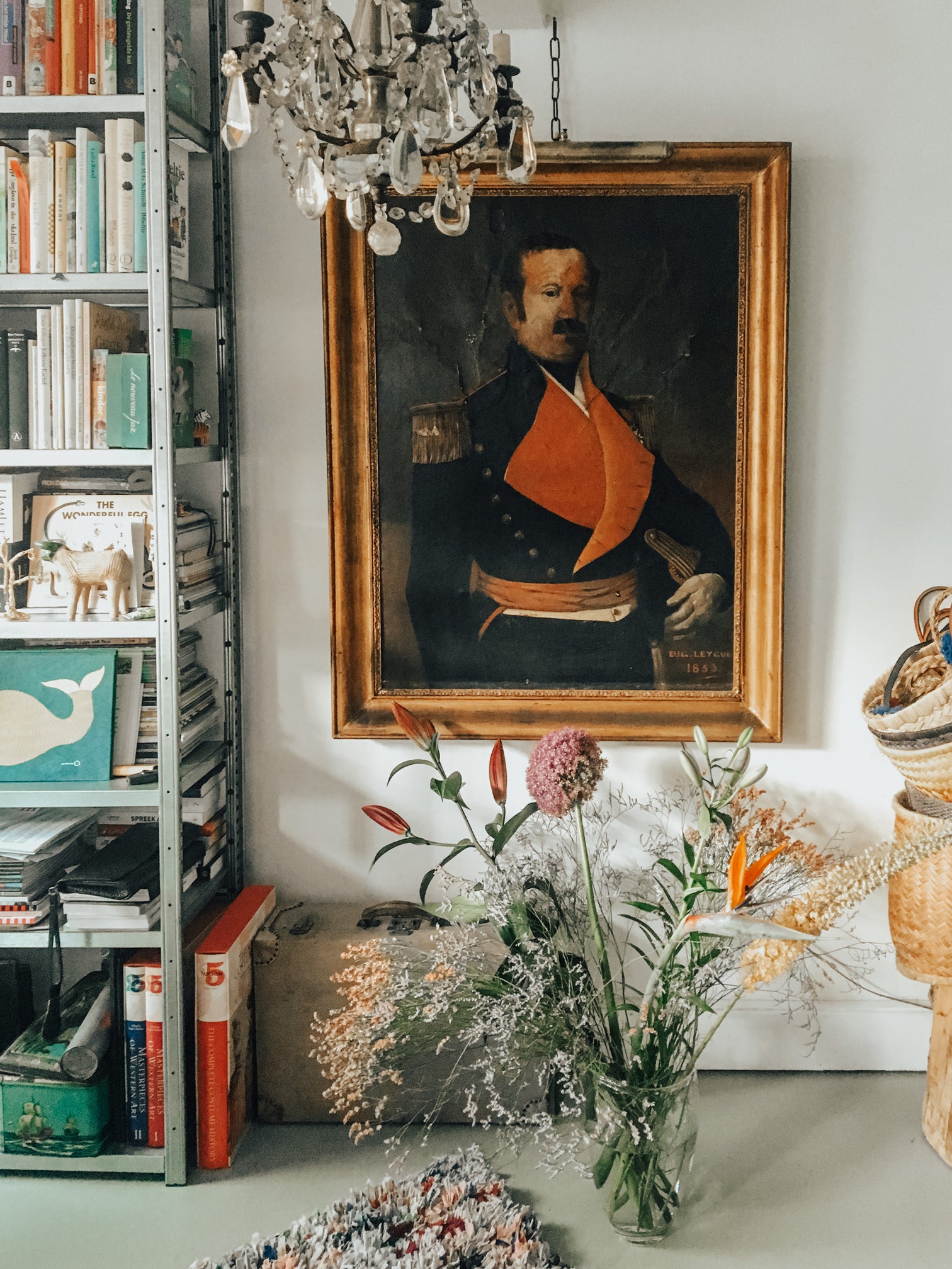 19th-century oil on canvas portrait of a French military man in a dark blue jacket with a red lapel, signed by Eug. Leycuf. Displayed in an interior with a grey-green floor, a bookcase filled with colorful books, and a vase of flowers placed in front. A small antique crystal chandelier hangs partially in front of the painting.