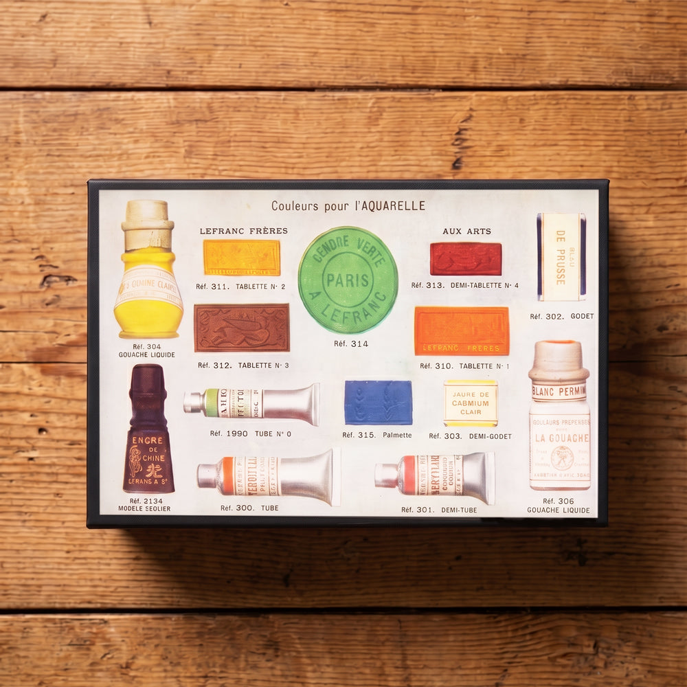 A large black rectangular matchbox featuring vintage-style illustrations and labels of watercolor paints and pigments, photographed on a rustic wooden background. Design by Archivist Gallery. Available at Wildschut Antiques & Decor.