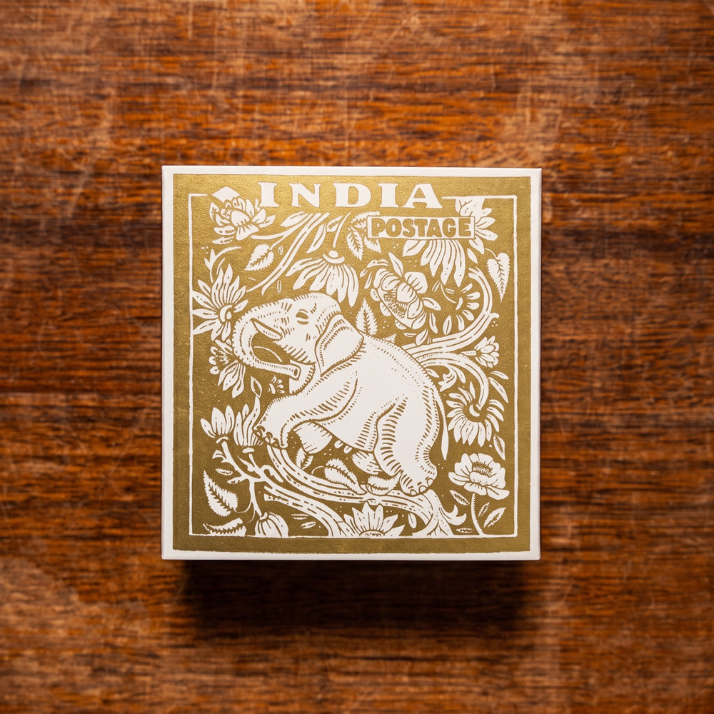 A vintage-style square matchbox featuring a postage stamp design with an elephant and floral illustrations with the text 'INDIA POSTAGE' on a rustic warm brown wooden surface. Design by Archivist Gallery. Available at Wildschut Antiques & Decor.