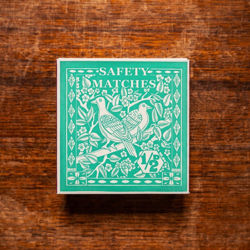 A square box of safety matches with a teal and white vintage-style bird illustration, placed on a rustic brown wooden surface. Design by Archivist Gallery. Available at Wildschut Antiques & Decor.