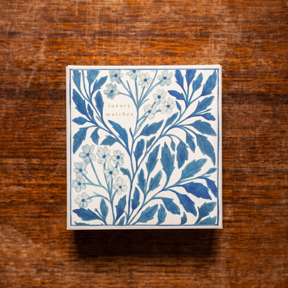A square box of luxury matches featuring a blue and white floral leaf pattern, placed on a rustic brown wooden surface. Design by Archivist Gallery. Available at Wildschut Antiques & Decor.