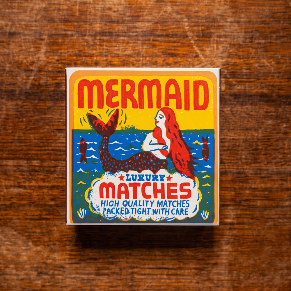 A square box of 'Mermaid Luxury Matches' featuring a colorful retro illustration of a mermaid on a yellow and blue background, placed on a rustic wooden surface.