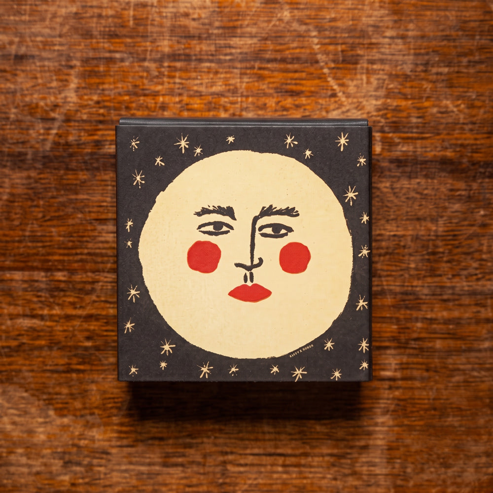 A square decorative matchbox with a minimalist face illustration on a dark background, placed on a wooden surface.