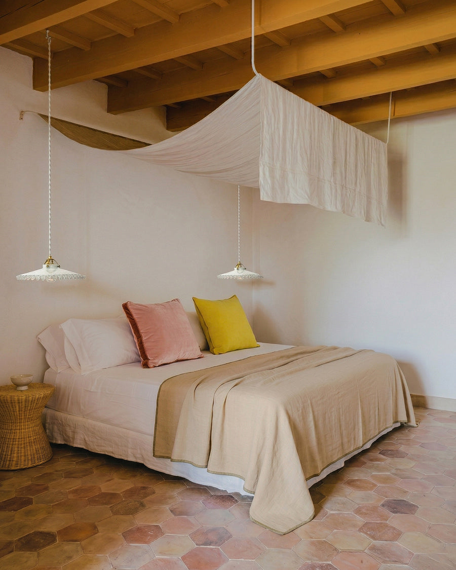 Bedroom with exposed wooden beams and terracotta tile flooring, centered around a low, linen-dressed bed in soft neutral tones. The bed is layered with warm beige textiles and accented by blush pink and ochre cushions. Above, a delicate, draped canopy floats like a suspended veil, flanked by two vintage-style pendant lights with scalloped glass shades. A woven wicker side table adds a natural touch, completing the serene, Mediterranean-inspired interior. Items all available via Wildschut Antiques.