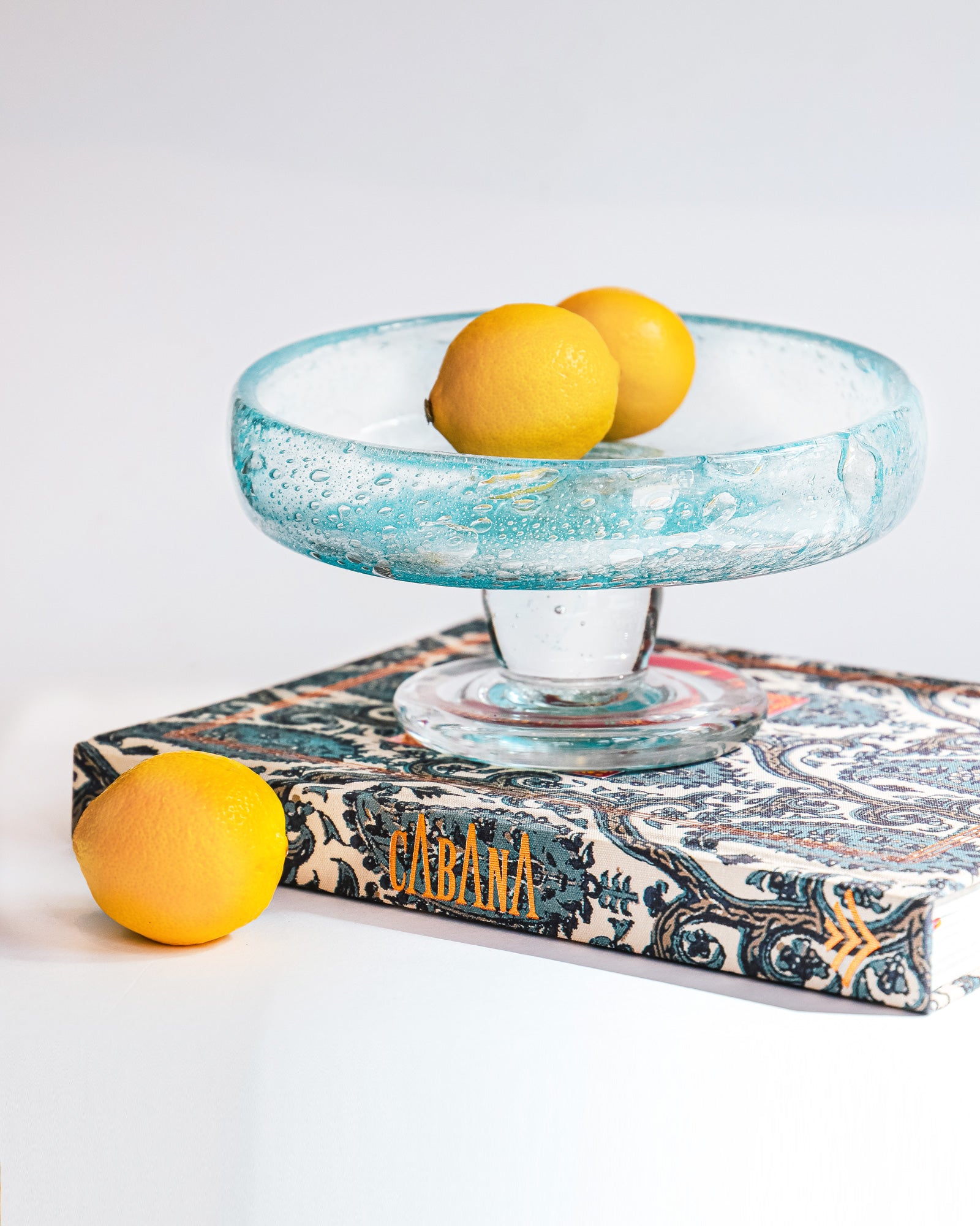Vintage round blue glass fruit bowl resembling a heavy glass droplet, likely by Biot, France, displayed with three lemons and a book by Cabana.