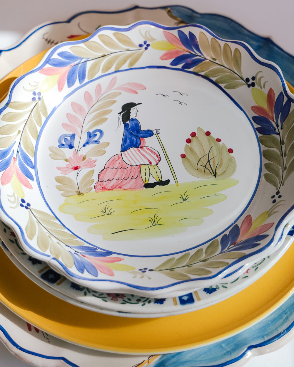 Stack of colorful ceramic plates topped with a vintage majolica plate featuring a hand-painted portrait of a man, scalloped edges, and floral motifs