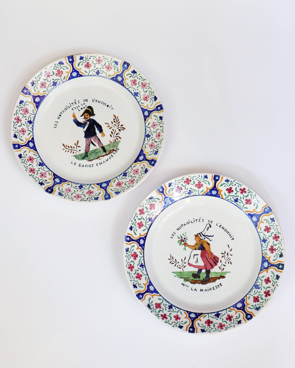 A set of two vintage Gien majolica ceramic plates with hand-painted depictions of a 'Garde Champére' and 'Madame La Mairesse' in a floral and colorful design