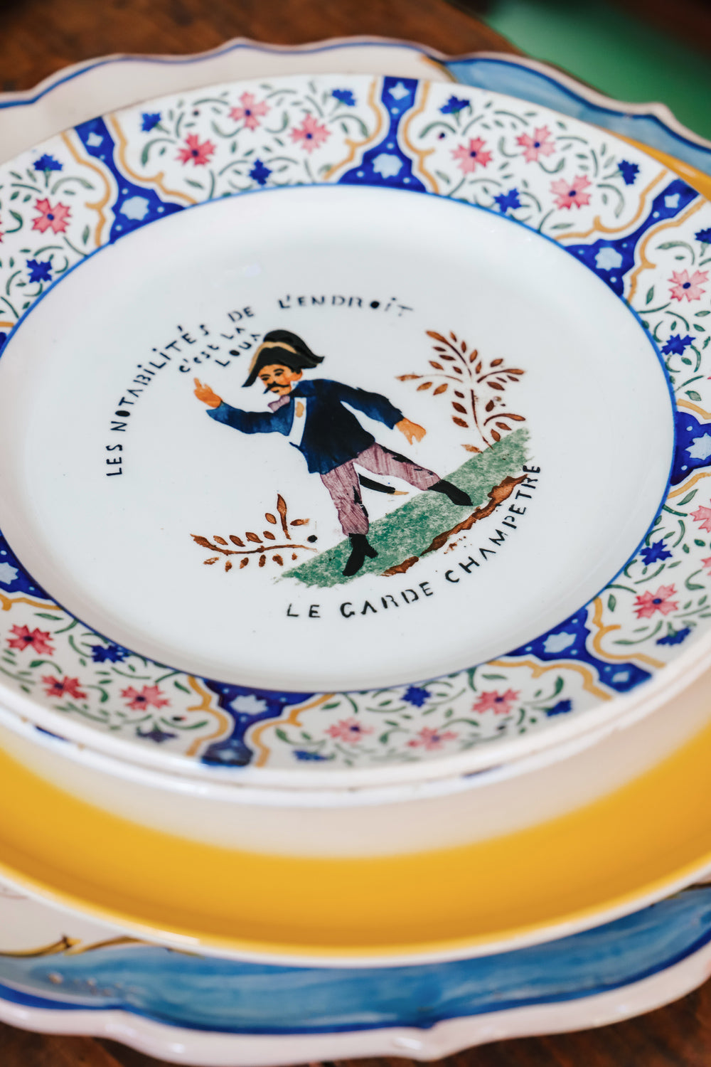 Stack of colorful vintage plates topped with a vintage Gien majolica plate, hand-painted with a vibrant floral design and a depiction of a *Garde Champêtre*.