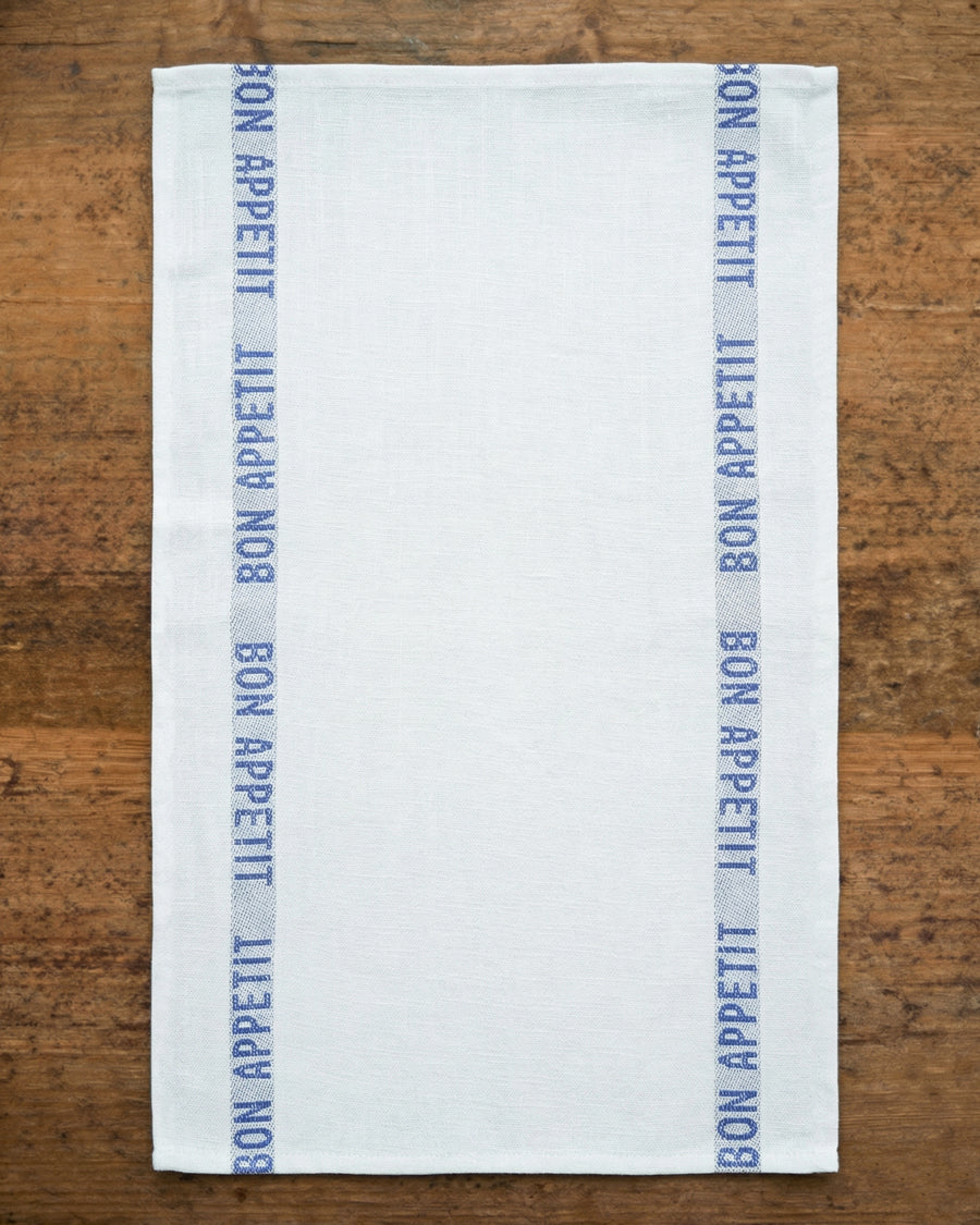 White linen towel with blue 'Bon Appetit' text on a wooden surface, by Charvet Editions available via Wildschut Antiques.