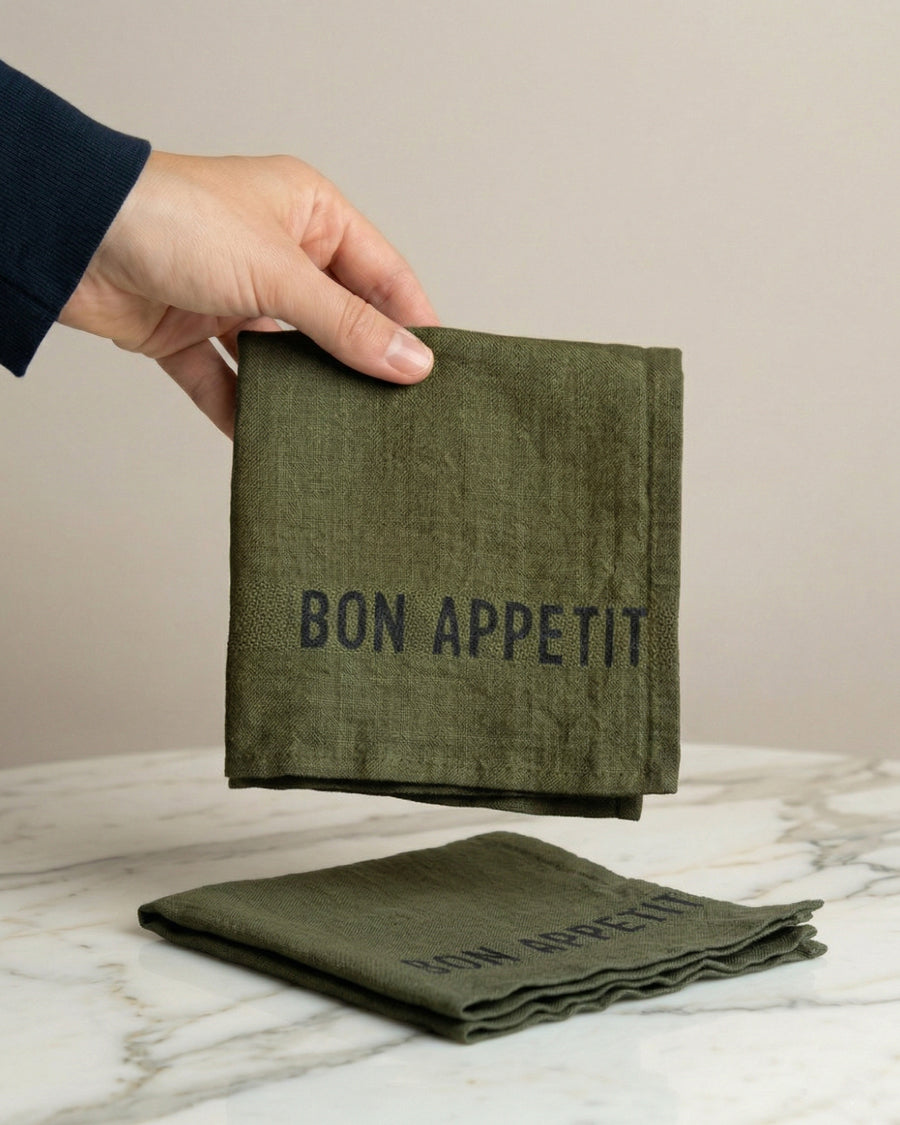 Green linen napkin with 'Bon Appetit' text held by a hand on a marble surface, by Charvet Editions available via Wildschut Antiques.
