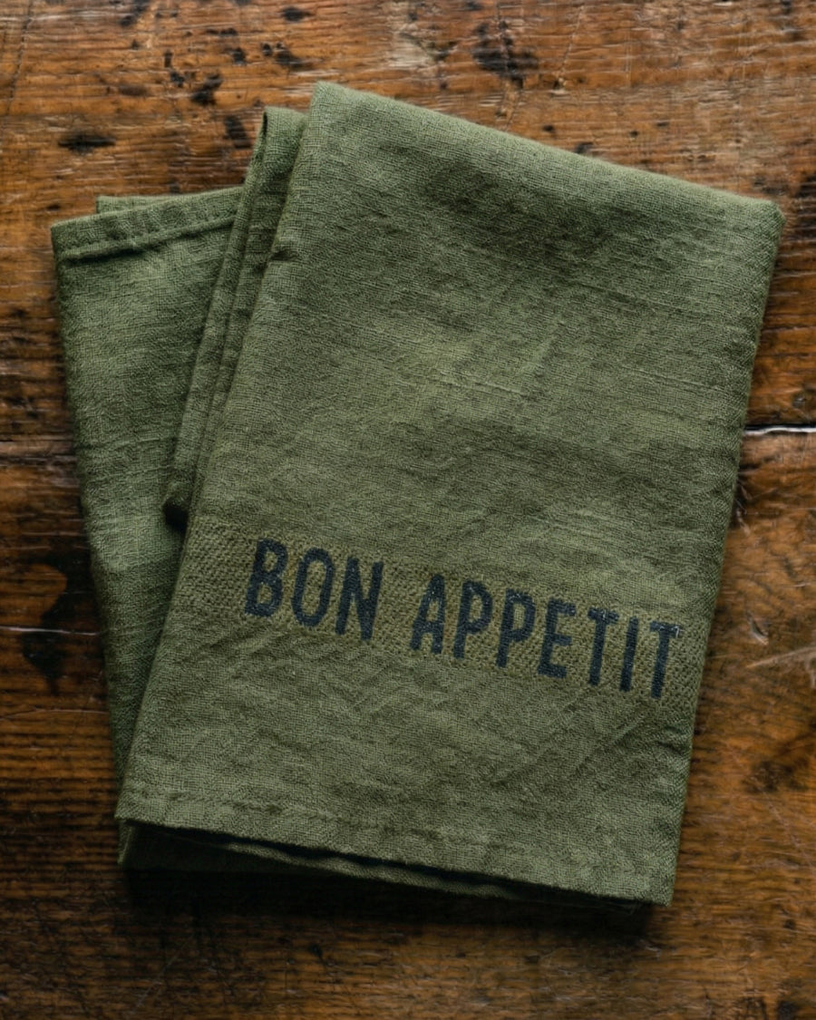 Green linen napkins with 'Bon Appetit' text on a wooden surface, by Charvet Editions available via Wildschut Antiques.