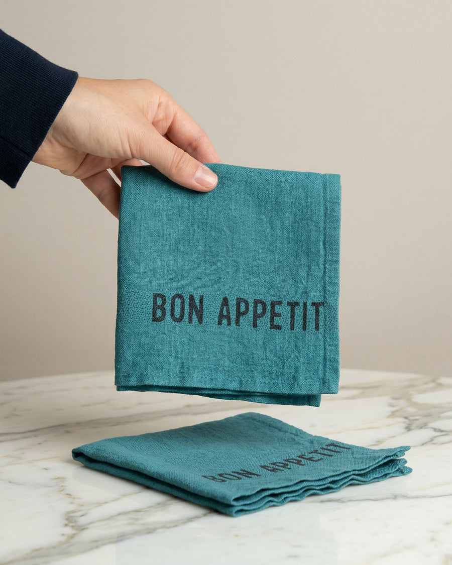 Teal linen napkin with 'Bon Appetit' text held by a hand on a marble surface, by Charvet Editions available via Wildschut Antiques.