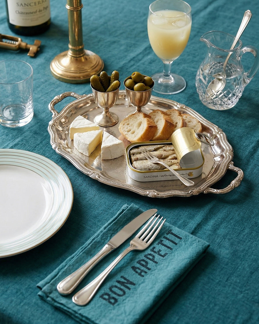 Table setting with a silver tray of food, glasses, and a 'Bon Appetit' linen napkin on a teal tablecloth, by Charvet Editions available via Wildschut Antiques.