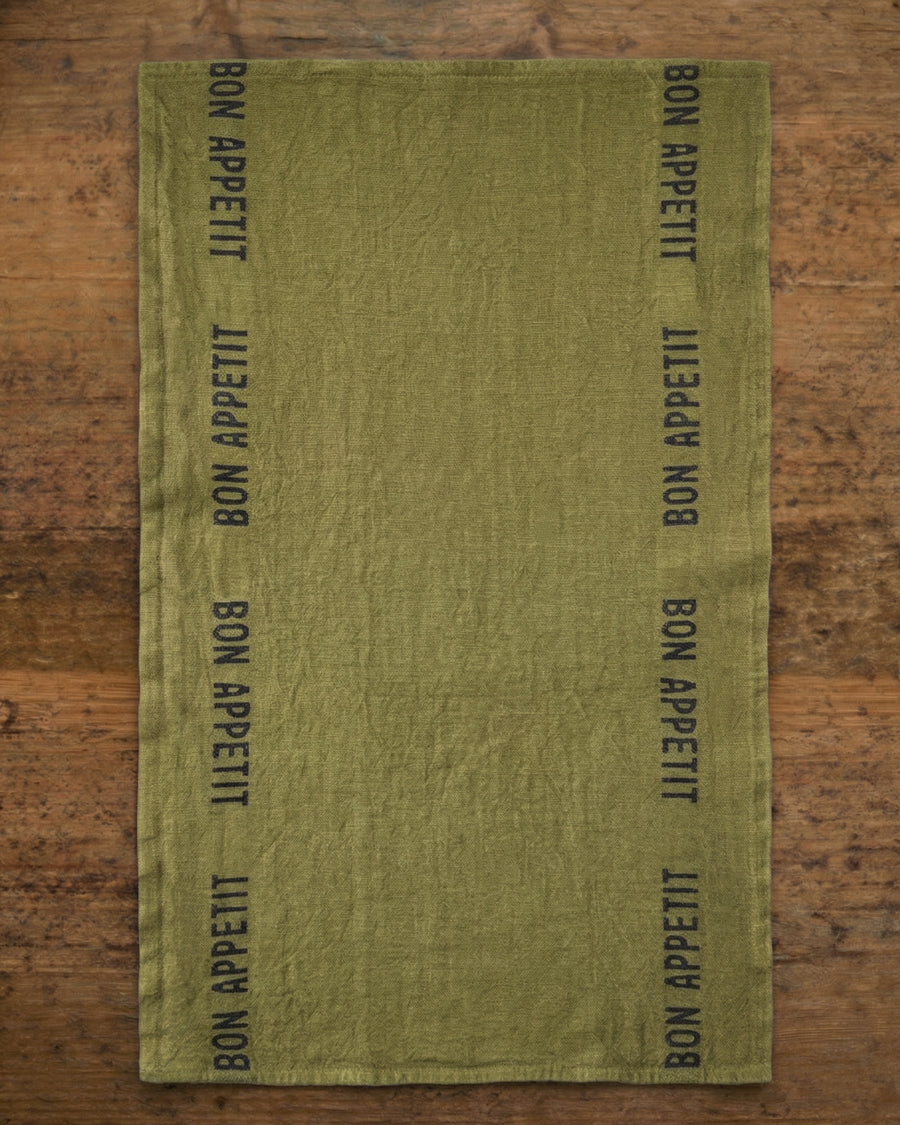 Olive green linen towel with 'Bon Appetit' text on a wooden surface, by Charvet Editions available via Wildschut Antiques.