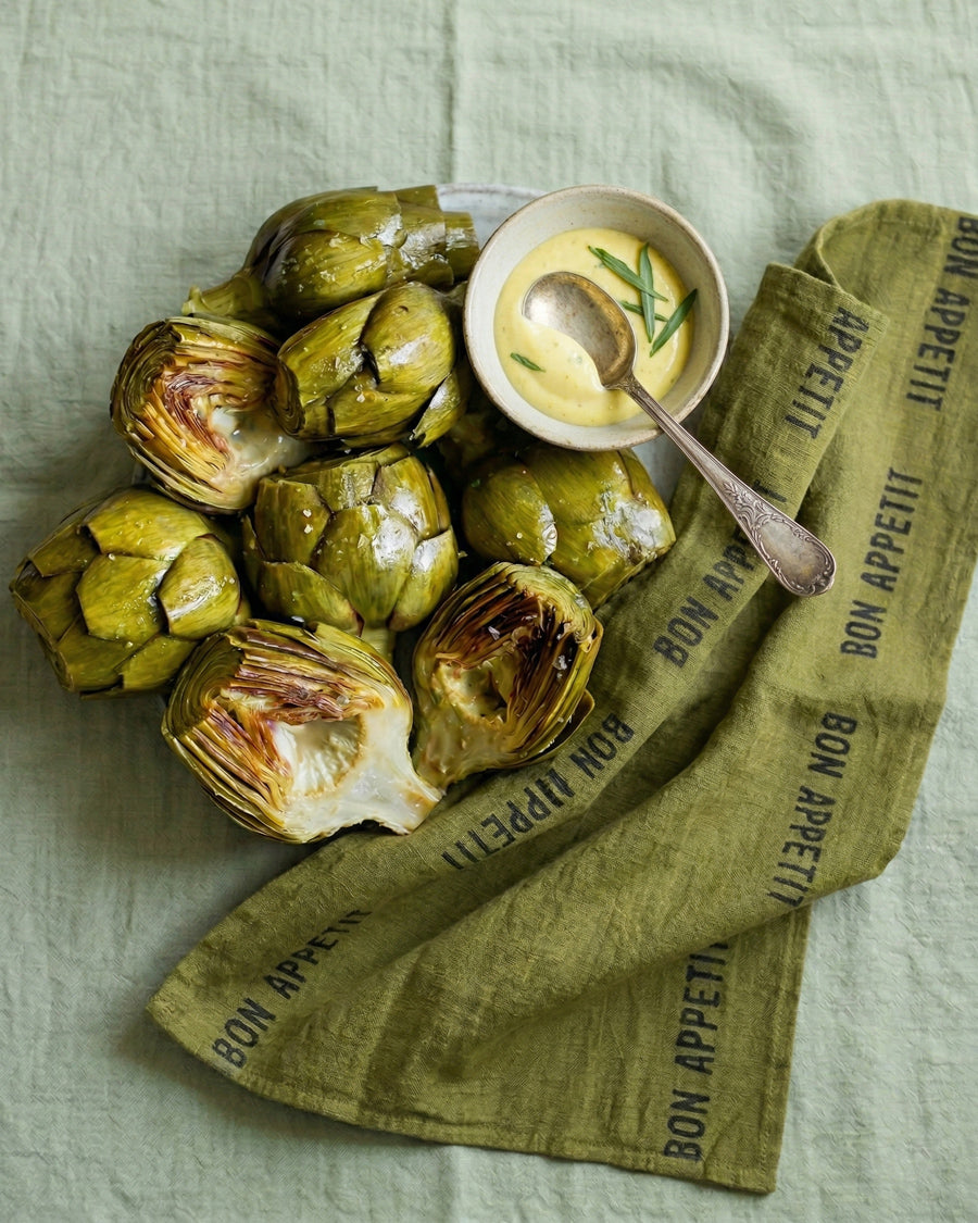 Artichokes with a bowl of dipping sauce next to a green linen towel with 'Bon Appetit' text by Charvet Editions available via Wildschut Antiques.