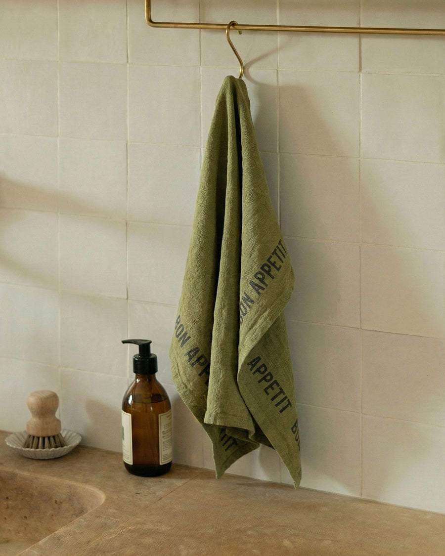Olive green linen towel with 'Bon Appetit' text hanging on a hook in a kitchen, by Charvet Editions available via Wildschut Antiques.