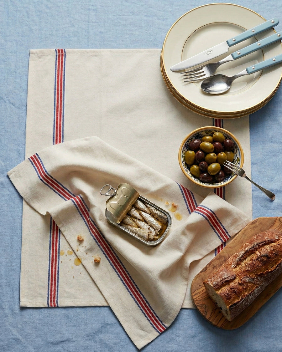 Table setting with bread, olives, and a can with sardines on a striped towel over a blue surface. Towels by Charvet Editions available via Wildschut Antiques.