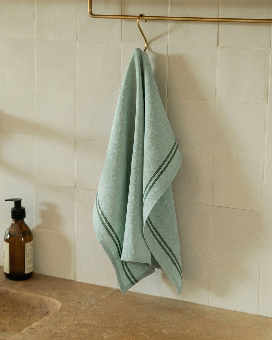 Mint green linen towel hanging on a gold hook against a tiled wall next to a bottle of soap in a kitchen. Towel by Charvet Editions available via Wildschut Antiques