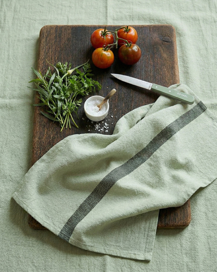 Wooden cutting board with herbs, tomatoes, a knife, and a light green towel on a light green surface. Linen towel by Charvet Editions available via Wildschut Antiques.