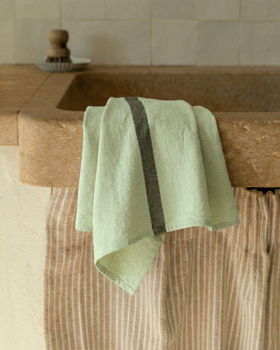 Soft pistache green linen towel hanging over a stone sink with a neutral tiled wall background, by Charvet Editions available via Wildschut Antiques.