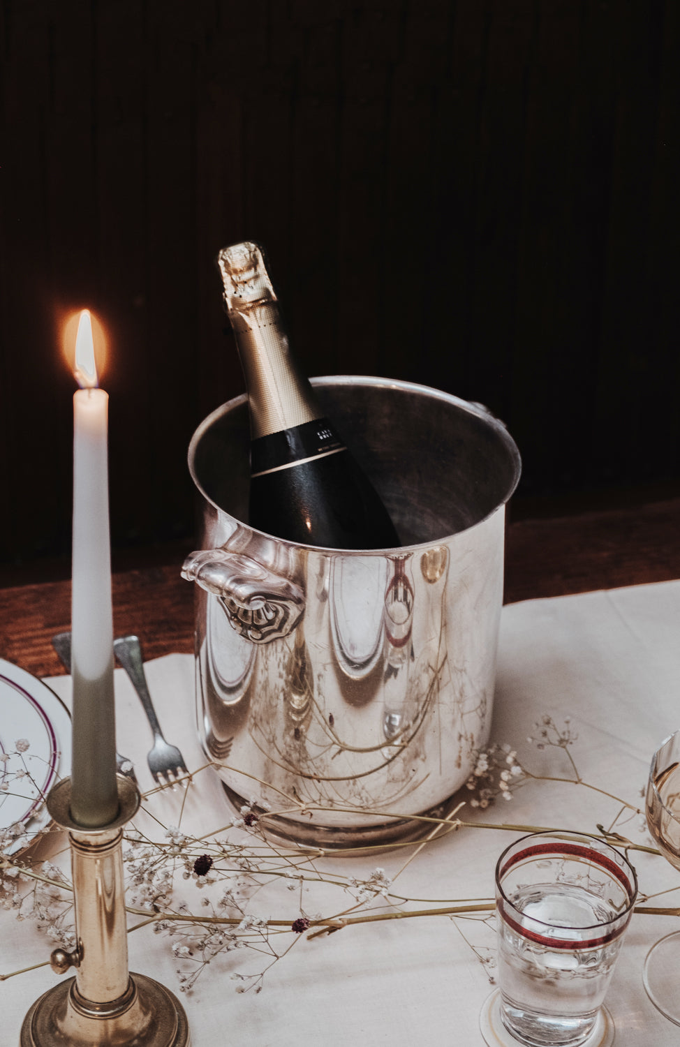 Elegant table setting featuring a bottle of champagne in a gleaming vintage Christofle Ormesson model cooler, placed beside a lit brass candleholder on a wooden table draped with white linen, at Wildschut Antiques.