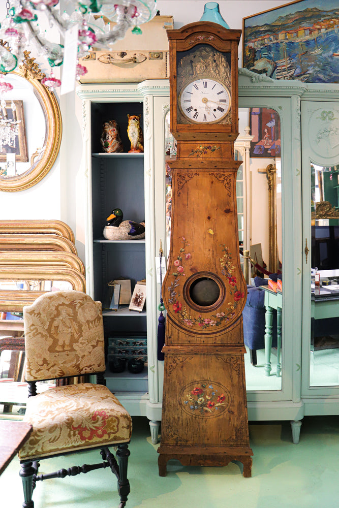 Antique French comtoise clock painted with floral details, positioned in front of a mint green mirror-door armoire in the Wildschut Antiques store.