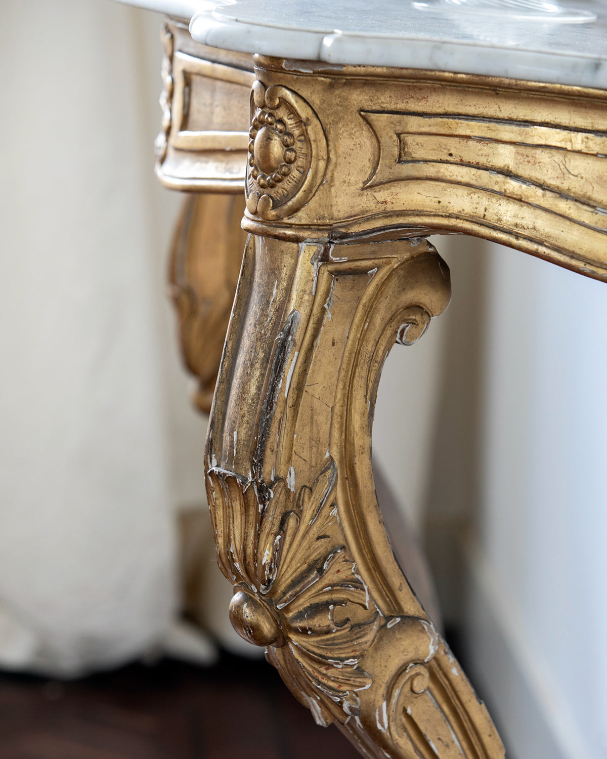 Close-up of a highly decorative antique French Louis XV style gilt wood console table with a white marble top, featuring intricate gilded designs and resting on carved legs. Showing some age appropriate wear and original patina.