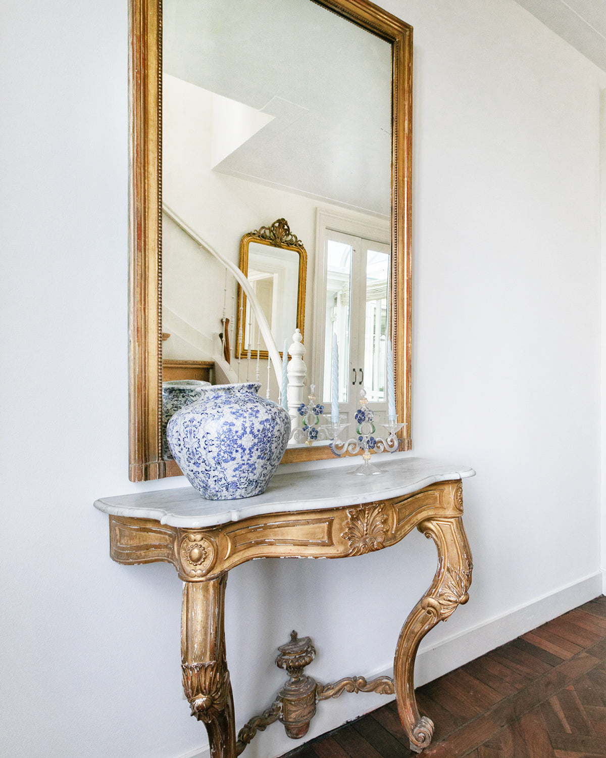 A highly decorative antique French Louis XV style gilt wood console table with a white marble top, featuring intricate gilded designs and resting on carved legs, displayed in a clean hallway with an antique golden mirror placed on top of the console