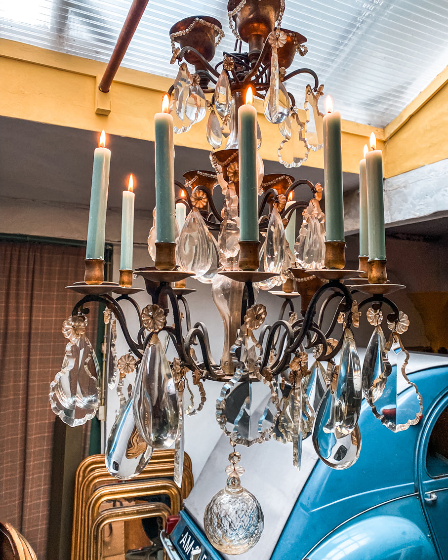Antique French crystal chandelier with cascading tiers, ornate bronze arms, real burning candles, and generous cut crystal drops and pendants. Finished with a faceted glass ball at the base for extra sparkle. Suspended from a yellow ceiling in the atmospheric warehouse of Wildschut Antiques with antique mirrors and a classic 2CV car in the background.