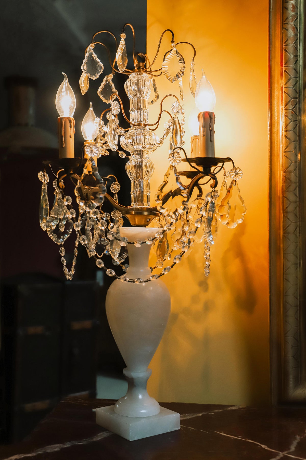 A 19th C French crystal chandelier-style table lamp with a white base, featuring a central candle holder and decorative crystal elements, against a yellow backdrop