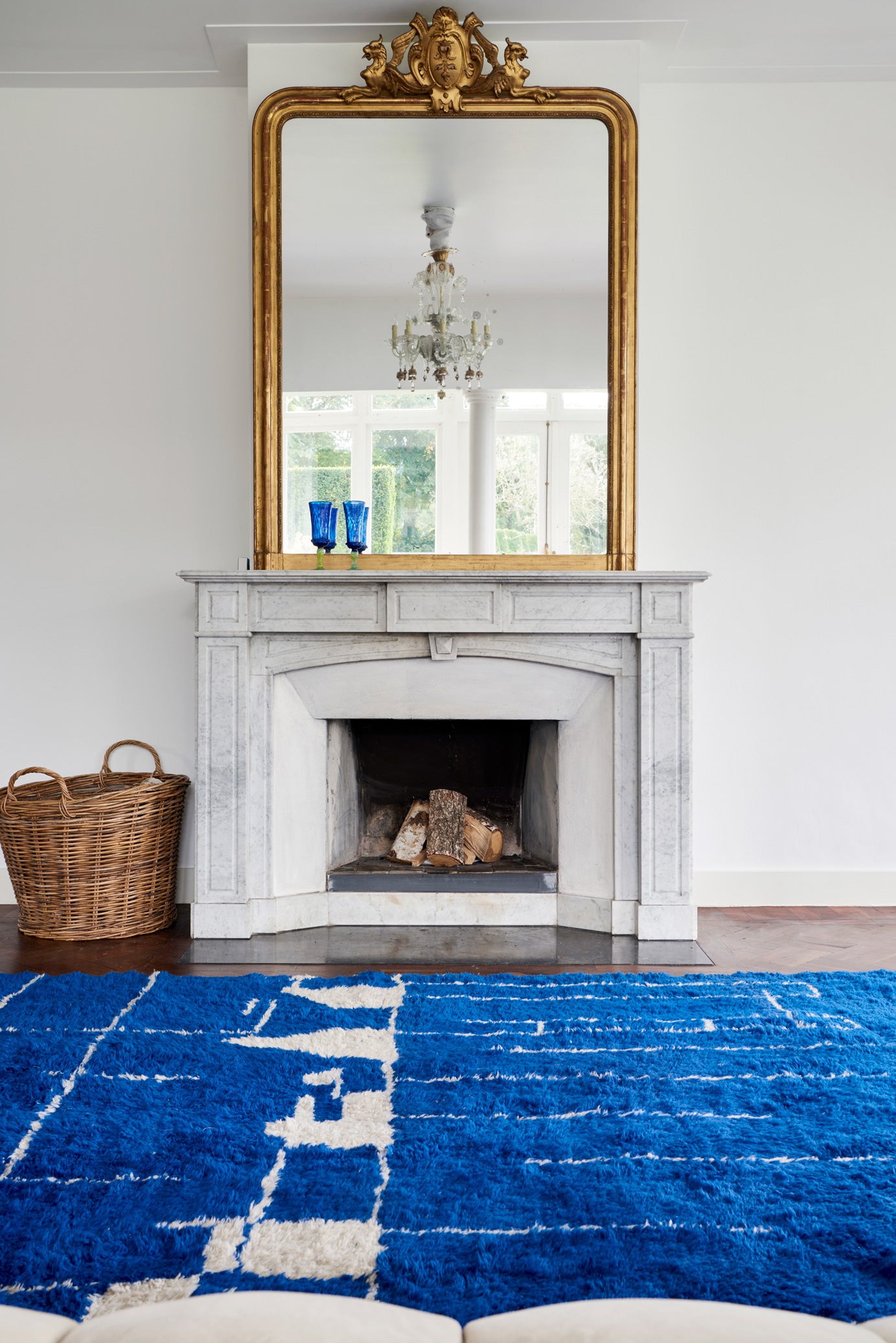An interior with a white marble fireplace topped with an antique gilt mirror reflecting a clear Venetian chandelier. A striking cobalt blue rug with white lines lies in front, with a woven wood basket positioned beside the hearth.