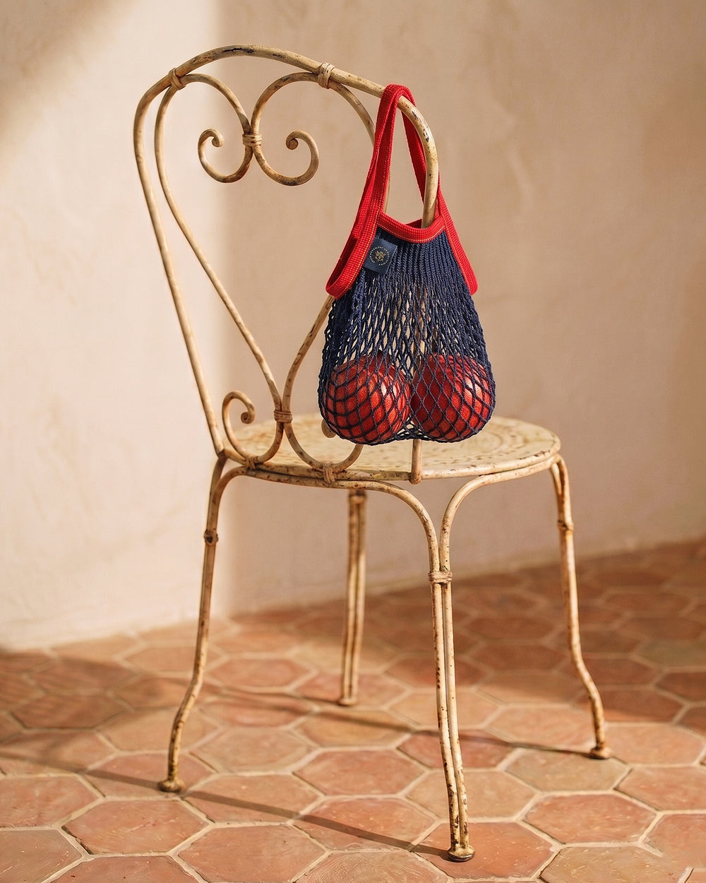 Red and blue mesh bag on a decorative vintage metal chair with a beige wall and tiled floor in the background. By Filt 1860, available at Wildschut Antiques.