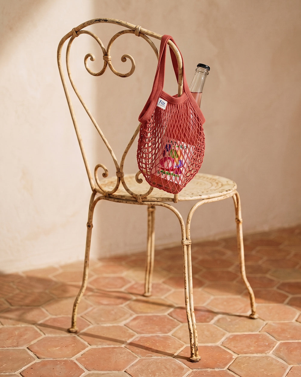 A red mesh reusable shopping bag with dual handles hangs from the back of a distressed gold metal chair, with a glass bottle tucked inside. The scene is set in a warm, rustic room with terracotta hexagonal floor tiles and a textured beige plaster wall. By Filt 1860, available at Wildschut Antiques.