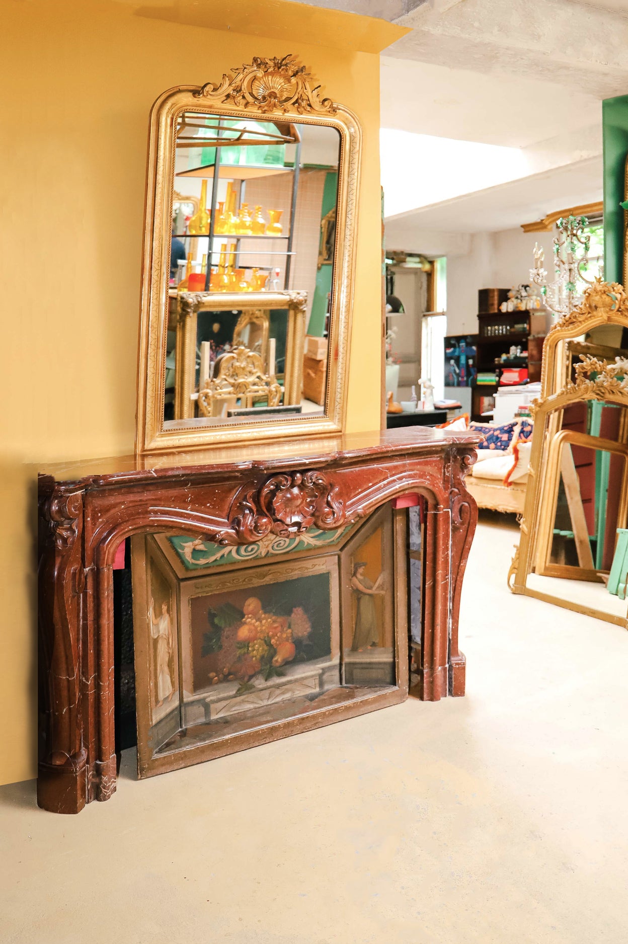 A grand antique French Rococo Louis XV fireplace mantel in richly veined red-brown marble, paired with an elegant painted firescreen featuring fruit and two graceful female figures. Surrounded by antique mirrors and a sofa layered with cushions, this striking vignette is set in the evocative warehouse of Wildschut Antiques.