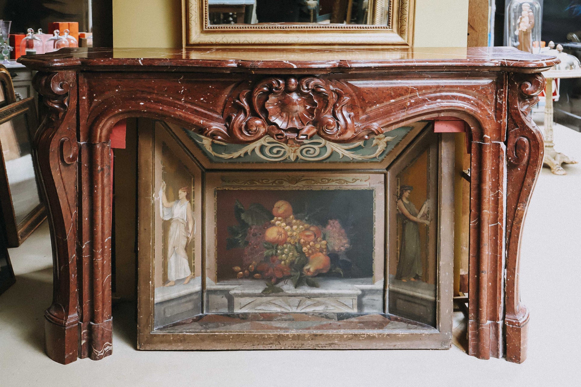 A grand antique French Rococo Louis XV fireplace mantel, crafted from richly veined red-brown marble. Displayed with an elegant painted firescreen adorned with fruits and two graceful female figures, all set in the atmospheric warehouse of Wildschut Antiques.