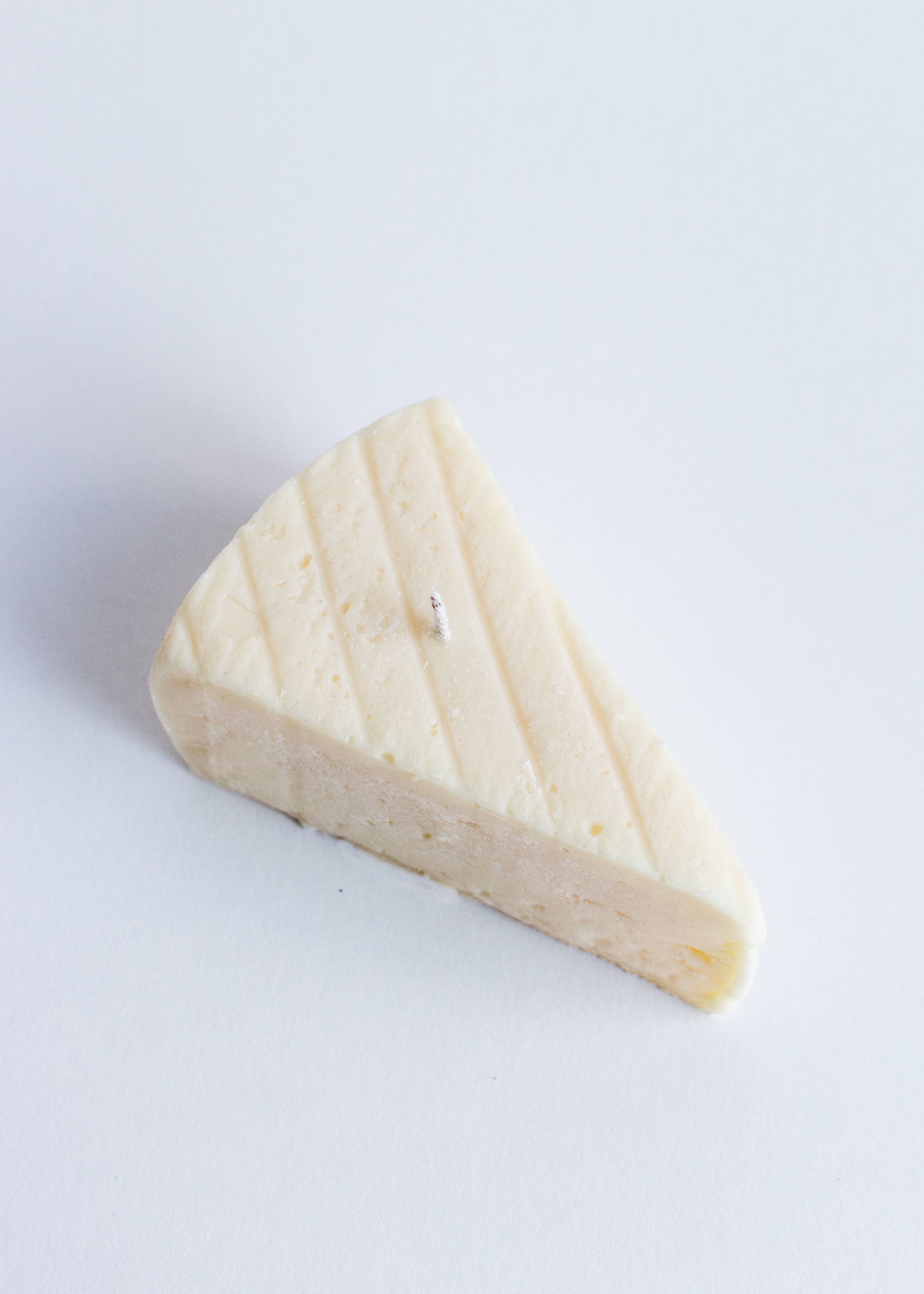 Triangle-shaped block of brie cheese on a white background. Fun fact: this is not a real piece of brie, it is a candle! Part of the Playful Food Candle Collection available via Wildschut Antiques Concept Store